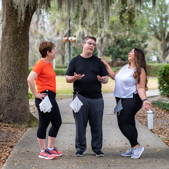 Three people standing on a sidewalk wearing Dry in Stride towels, in a park-like setting, engaged in conversation.