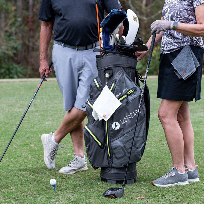Two people on a golf course with a golf bag and clubs. One is wearing a Dry in Stride towel and one has it clipped to the golf bag.